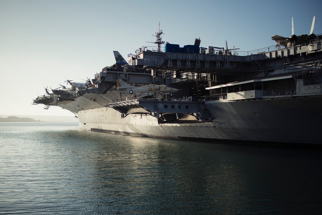 A large warship docked in calm waters under a clear sky, showcasing its structure and military aircraft on deck.